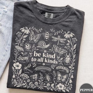 Pode incluir: Uma t-shirt cinza-carvão com uma estampa gráfica branca de vários insetos e flores rodeando o texto "be kind to all kinds". O design inclui ilustrações de borboleta, abelha, libélula, caracol, joaninha e outras pequenas criaturas.
