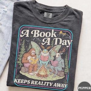 May include: A charcoal gray t-shirt featuring a graphic of Bigfoot, a moth creature, and a deer-headed figure sitting around a campfire reading books. The text reads A Book A Day Keeps Reality Away. The shirt has a crew neck and short sleeves.
