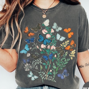 May include: Dark gray t-shirt featuring a colorful butterfly and floral design. The graphic includes butterflies in shades of blue, orange, and white, with pink flowers and green foliage. The shirt has a vintage, worn look.