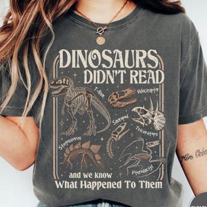 May include: Dark gray t-shirt with a dinosaur-themed graphic. The design features dinosaur skeletons and skulls, with the text "DINOSAURS DIDN'T READ and we know What Happened To Them". The shirt is a comfortable, casual wear.
