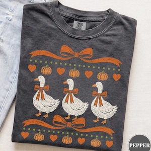 May include: A charcoal grey t-shirt featuring a graphic print of three white geese wearing orange bows. The design includes orange ribbons, small orange hearts, and orange pumpkins arranged in rows. The shirt has a crew neckline and a Comfort Colors label.