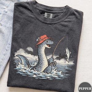 May include: A charcoal gray t-shirt featuring a graphic of a blue sea creature wearing a red fishing hat and holding a fishing rod with a hooked fish. The shirt has a crew neckline and a Comfort Colors brand label printed on the inside collar.