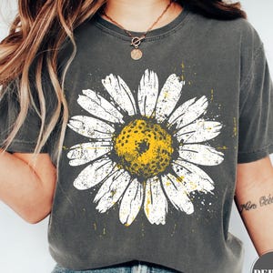 Puede incluir: Camiseta gris oscuro con una gran margarita blanca y centro amarillo. El dise&ntilde;o de la margarita tiene un aspecto envejecido y vintage. La camiseta es de cuello redondo y tiene mangas cortas. La palabra "Pepper" est&aacute; impresa en la parte inferior derecha.