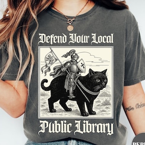 May include: Dark gray t-shirt with a graphic of a knight riding a black cat, holding a flag. The text "Defend Your Local Public Library" is above and below the graphic. The shirt is a comfortable, casual style.