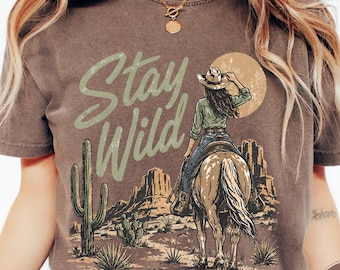 May include: Brown t-shirt with a desert-themed graphic. The design features a cowgirl on horseback, cacti, and the words "Stay Wild" in a light green font. The shirt has a vintage, worn look.