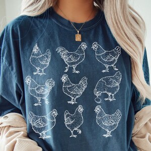 May include: Navy blue t-shirt featuring a white illustration of nine chickens. The shirt has a crew neck and short sleeves. The words "NAVY BLUE" are in a white circle in the lower right corner. The shirt is made of a soft material.