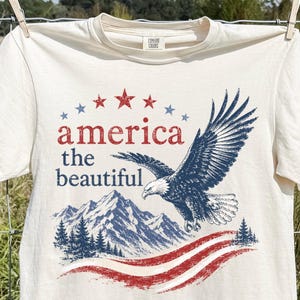 May include: An ivory short-sleeve t-shirt featuring a graphic print of a bald eagle in flight above mountain peaks and pine trees. The text reads "america the beautiful" in red and blue, accompanied by red and blue stars and wavy stripes.