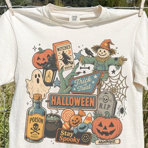 May include: An ivory short-sleeve t-shirt featuring a colourful Halloween-themed graphic print. The design includes a scarecrow, a ghost, a pumpkin, a poison bottle, a cauldron, a tombstone, and text reading Witches Brew, Trick or Treat, Halloween, and Stay Spooky.