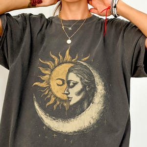 May include: Dark gray t-shirt featuring a sun and moon graphic with a woman's face. The design is in shades of gold and white. The shirt has short sleeves and a relaxed fit.