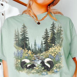 May include: A light green t-shirt featuring a nature scene with two skunks, a stream, and a forest. The skunks are black and white, and the forest has tall green trees. The shirt has the word "BAY" printed on it.