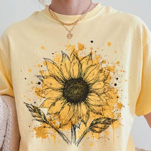 Könnte beinhalten: Ein hellgelbes Kurzarm-T-Shirt mit einer großen, detaillierten Sonnenblumen-Illustration im schwarzen Tusche-Stil auf der Brust. Das Design ist mit leuchtend gelben Farbspritzern akzentuiert. Ein kleines kreisförmiges Etikett trägt die Aufschrift BANANA.