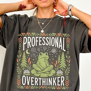 May include: A charcoal grey short-sleeve t-shirt featuring a graphic print of a green frog sitting on a lily pad. The text reads "PROFESSIONAL OVERTHINKER" in white, surrounded by illustrations of pine trees, cattails, and geometric patterns.