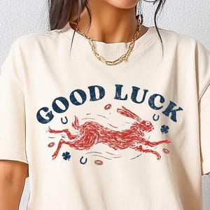May include: Ivory-colored t-shirt with the words "GOOD LUCK" in navy blue, featuring a red rabbit graphic. The design includes horseshoes and shamrocks. A casual, comfortable tee.