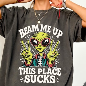 May include: Dark gray t-shirt with a graphic of an alien wearing sunglasses and making a peace sign. The text on the shirt reads "Beam Me Up This Place Sucks."