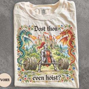 Könnte beinhalten: Ein elfenbeinfarbenes T-Shirt mit einem Grafikdruck eines Ritters in voller Rüstung, der eine Langhantel mit Steingewichten hebt. Im Hintergrund steht eine Burg, flankiert von einem blauen und einem roten Drachen. Der Text lautet: Dost thou even hoist?