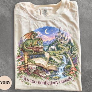 May include: An ivory t-shirt featuring a graphic print of a green dragon perched on a stack of books, a castle, a unicorn, a waterfall, flowers, a candle, and glasses. The text "it's too nonfictiony outside" is printed in dark purple at the bottom.