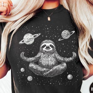 May include: Black t-shirt with a cosmic design featuring a meditating sloth, planets, and stars. The sloth is depicted in a yoga pose. The word "BLACK" is in a circle on the bottom right.
