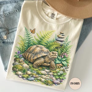 Puede incluir: Una camiseta color marfil con un gráfico de una tortuga rodeada de helechos verdes, flores pequeñas, una mariposa y piedras apiladas. La prenda se combina con vaqueros azul claro, un sombrero beige de ala ancha y un collar de plata.