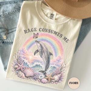 May include: An ivory t-shirt featuring a graphic of a dolphin jumping over ocean waves with a rainbow, a butterfly, and seashells. The text "RAGE CONSUMES ME" is printed above the graphic. Also includes light blue denim jeans and a tan hat.