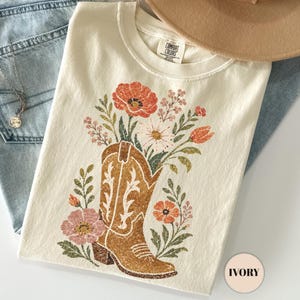 以下が含まれることがあります： アイボリーのTシャツには、オレンジ、ピンク、白の花で満たされた茶色のカウボーイブーツのグラフィックプリントが施されています。このシャツは、ライトブルーのデニムジーンズ、ベージュのつば広帽子、小さなチャームが付いた繊細なシルバーチェーンネックレスと組み合わされています。