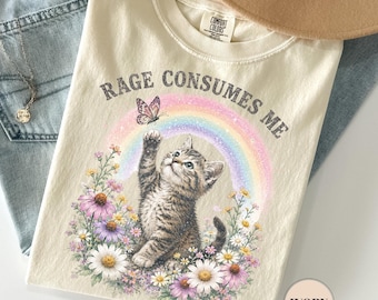 May include: Ivory-colored t-shirt with the text "RAGE CONSUMES ME" and a graphic of a kitten reaching for a butterfly in front of a rainbow. The shirt is paired with light blue jeans, a necklace, and a tan hat.