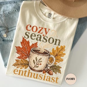 May include: An ivory t-shirt featuring the text "cozy season enthusiast" in orange and brown, a graphic of a steaming mug, autumn leaves, and acorns. The shirt is layered with light blue denim jeans and a tan wide-brimmed felt hat.