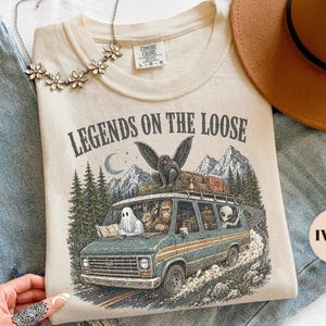 May include: An ivory t-shirt featuring a graphic of a van with a ghost, Bigfoot, and an alien, topped with a moth creature. The text reads LEGENDS ON THE LOOSE. The design includes mountains, pine trees, and a crescent moon in a vintage style.