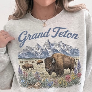 May include: A light gray crewneck sweatshirt featuring a graphic print of the Grand Teton mountains, a large bison in the foreground, smaller bison in the distance, and colorful wildflowers. The text Grand Teton is printed in blue above the mountains.