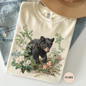 Puede incluir: Camiseta de color marfil con un diseño de osezno negro rodeado de flores y follaje. Se combina con vaqueros azul claro, un sombrero beige y un collar plateado. La etiqueta de la camiseta dice "COMFORT COLORS".
