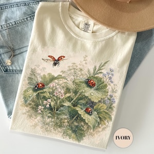 May include: Ivory-colored Comfort Colors t-shirt featuring a botanical illustration of ladybugs on leaves. The shirt is paired with light blue jeans, a tan hat, and a delicate necklace. The shirt's design includes the word "IVORY".
