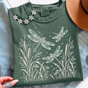 Op de afbeelding: Een mosgroen t-shirt met een witte grafische print van twee libellen die over lisdodden en wilde bloemen vliegen, met kleine fonkelende sterren. Het shirt wordt gecombineerd met een blauwe spijkerbroek, een bruine vilten hoed en een zilveren ketting.