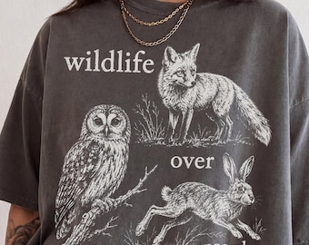 May include: A charcoal gray short-sleeve t-shirt featuring a white graphic print of an owl, a fox, and a rabbit. The text "wildlife over people" is printed in white lettering across the front. The shirt has a crew neckline and a relaxed fit.