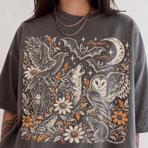 Może przedstawiać: Ciemnoszary t-shirt z krótkim rękawem z nadrukiem graficznym przedstawiającym kruka, wyjącego wilka, sowę, nietoperze, pająka w sieci, żabę, szczura, kwiaty i półksiężyc. Koszulka zawiera okrągłą naszywkę z napisem PEPPER.