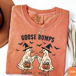 May include: An orange Comfort Colors t-shirt with the words "GOOSE BUMPS" and a graphic of two cartoon geese wearing witch hats and holding jack-o'-lanterns. Bats are flying above the geese. A gold necklace is draped across the shirt.