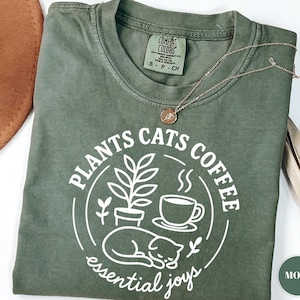 Op de afbeelding: Een mosgroen t-shirt met de woorden "PLANTS CATS COFFEE" in een cirkelvormig ontwerp, met een illustratie van een plant, kat en koffiekopje. De zin "essential joys" staat eronder. Een gouden ketting ligt over het shirt.