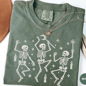 May include: Moss green Comfort Colors t-shirt featuring a whimsical design of three dancing skeletons with candles and stars. The skeletons are holding a crescent moon, a star wand, and a candle. A gold necklace is draped across the shirt.