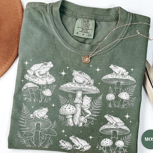 May include: Moss green t-shirt with a white graphic of frogs on mushrooms. The design includes various mushroom shapes and ferns. The shirt has a crew neck and short sleeves. A gold necklace is draped across the shirt.