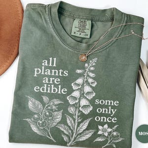 以下が含まれることがあります： モスグリーンのクルーネックTシャツ。フォックスグローブとベリーの白い植物画が描かれています。テキストは「all plants are edible some only once」。襟元にはComfort Colorsのラベルとゴールドのネックレスが付いています。