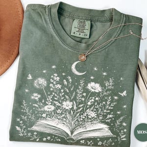Op de afbeelding: Een mosgroen t-shirt met korte mouwen en een witte grafische print van een open boek met wilde bloemen, vlinders en een maansikkel die uit de pagina's oprijst. Het shirt heeft een ronde hals en een Comfort Colors label in de nek.