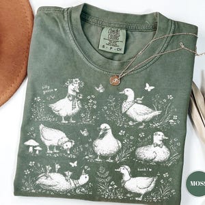Peut inclure: T-shirt vert mousse avec une illustration fantaisiste de canards dans diverses poses, ornés d'éléments floraux et de papillons. L'expression "silly goose" est imprimée sur le t-shirt. Un collier en or avec un pendentif est posé sur le t-shirt.