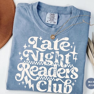 Könnte beinhalten: Ein blaues Comfort Colors T-Shirt mit dem Text "Late Night Readers Club" in weißer Retro-Schrift. Das Shirt hat einen Rundhalsausschnitt und kurze Ärmel. Eine goldene Halskette mit Anhänger liegt über dem Shirt.