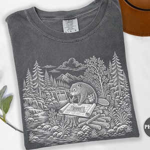 May include: A charcoal grey t-shirt with a white graphic print of a beaver working on a blueprint at a dam site. The shirt features a crew neckline and a Comfort Colors label. The graphic depicts a forest, mountains, and a waterfall.