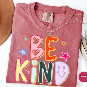 Könnte beinhalten: Ein altrosa T-Shirt mit Rundhalsausschnitt und einem Comfort Colors Etikett. Der Aufdruck zeigt den Text BE KIND in bunten, fetten Buchstaben mit goldenen Glitzerumrandungen, akzentuiert durch kleine Sterne, Blumen und ein lila Smiley-Gesicht.