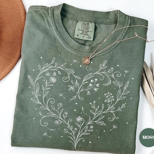 May include: A moss green t-shirt with a white floral heart design. The shirt has a crew neck and short sleeves. A gold necklace with a pendant rests on the shirt. The tag reads "Comfort Colors".