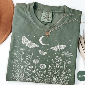 May include: A moss green short-sleeve t-shirt featuring a white graphic print of three moths, a crescent moon, stars, and various wildflowers. The shirt has a crew neckline and a Comfort Colors brand label inside the collar. A thin gold chain necklace.