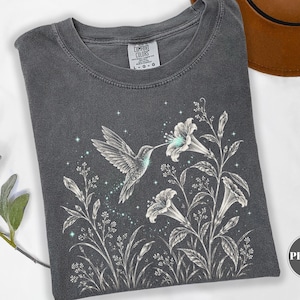 Pode incluir: Uma t-shirt cinzento-carvão com uma ilustração de linhas brancas de um beija-flor a pairar perto de flores em forma de trombeta. O design inclui pequenos detalhes de estrelas e um toque de cor azul-claro na flor. A t-shirt tem uma etiqueta Comfort Colors.