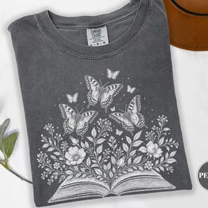Puede incluir: Una camiseta gris carbón con un gráfico de líneas blancas de un libro abierto del que brotan flores y hojas, rodeado de varias mariposas. La camiseta tiene cuello redondo y una etiqueta de la marca Comfort Colors en el cuello.