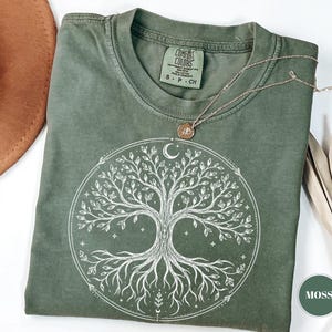 May include: A moss green short-sleeve t-shirt featuring a white circular graphic of a tree with intricate roots and branches, a crescent moon, and small stars. The inner neck label reads Comfort Colors, and a thin gold-tone necklace rests on the fabric.