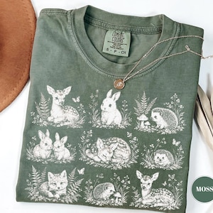 May include: A moss green t-shirt featuring a woodland animal design. The graphic includes deer, rabbits, hedgehogs, and a fox, surrounded by ferns and flowers. A gold necklace with a pendant rests on the shirt. The shirt is labeled "Comfort Colors".