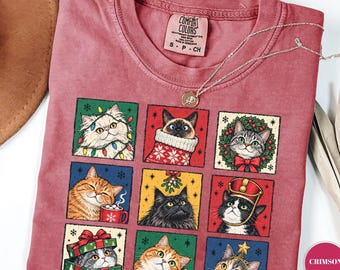 May include: A crimson t-shirt featuring a grid of nine illustrated cats in festive holiday settings, including cats with Christmas lights, a stocking, a wreath, a mug, mistletoe, a gift box, and ornaments. The shirt has a Comfort Colors label.